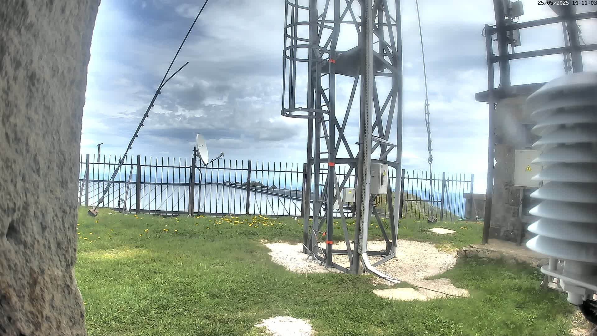 A metal communications tower stands on a grassy area with a cloudy sky, a satellite dish, and a fence in the background.