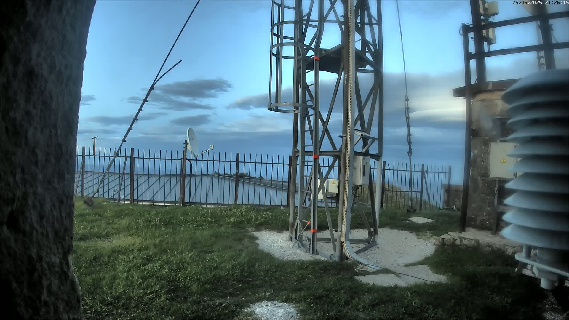 A metal tower with a staircase stands near a fence and grassy area under a partly cloudy sky.