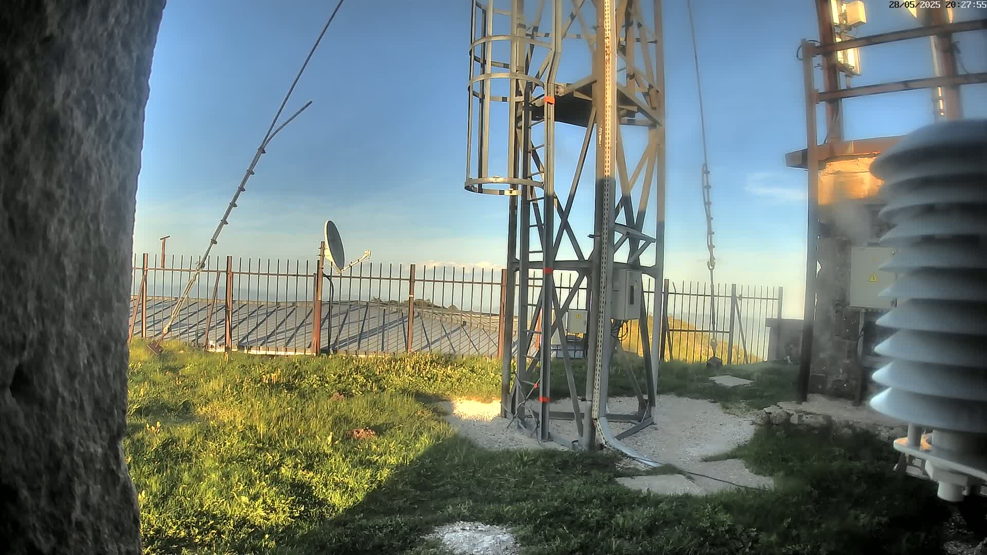 A metal communications tower with a satellite dish and weather station stands on a grassy hilltop under a clear blue sky.