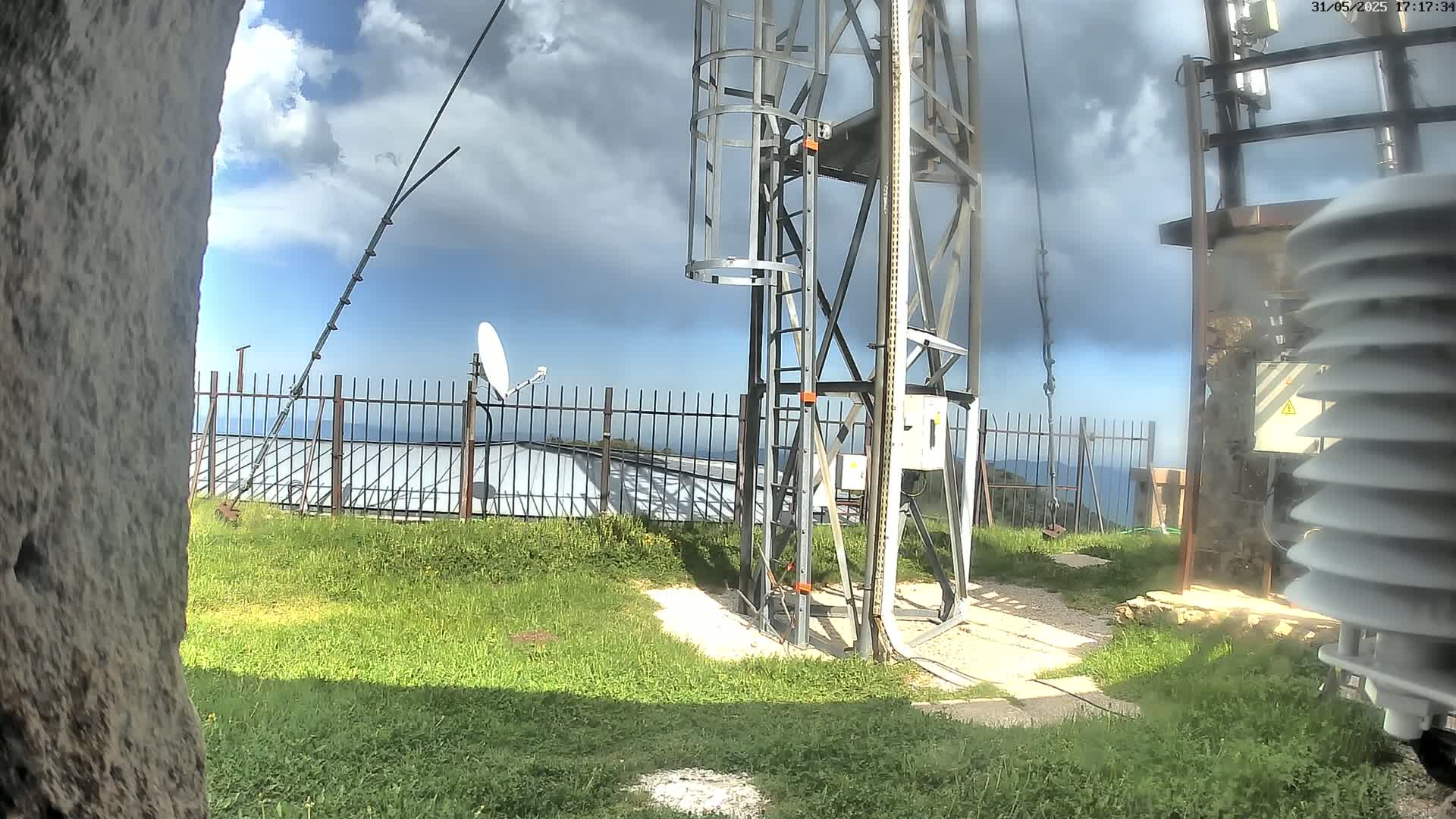 A metal communications tower with a ladder and attached equipment stands on grassy ground overlooking a fence and a distant body of water under a partly cloudy sky.