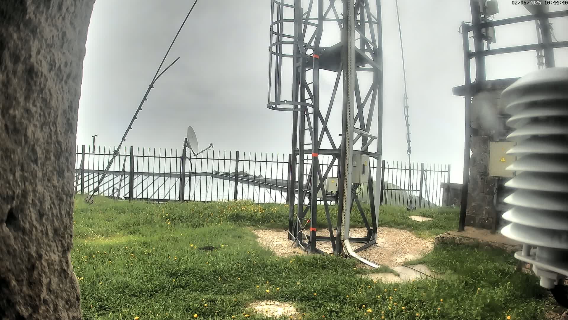 A metal tower with a satellite dish and other equipment stands behind a fence in a grassy area on an overcast day.