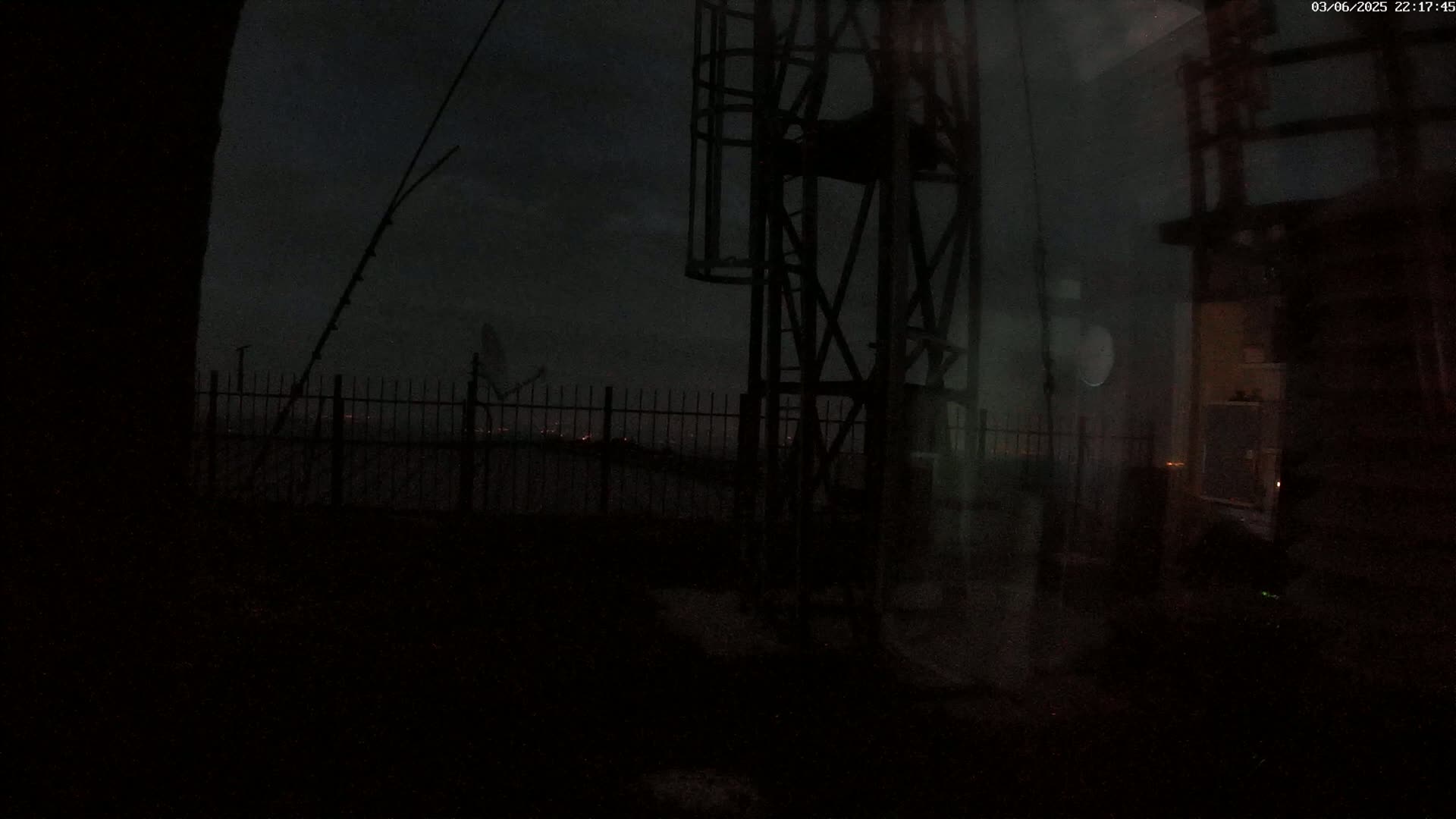 A dark, nighttime scene shows a metal structure next to a fence overlooking a distant, dimly lit cityscape under a cloudy sky.