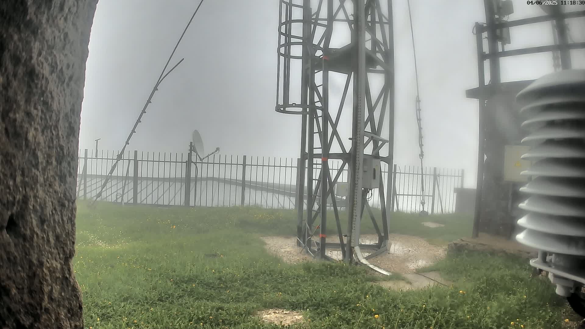 A metal tower with a ladder and various equipment is partially visible through a thick fog, surrounded by a grassy area with a short fence.
