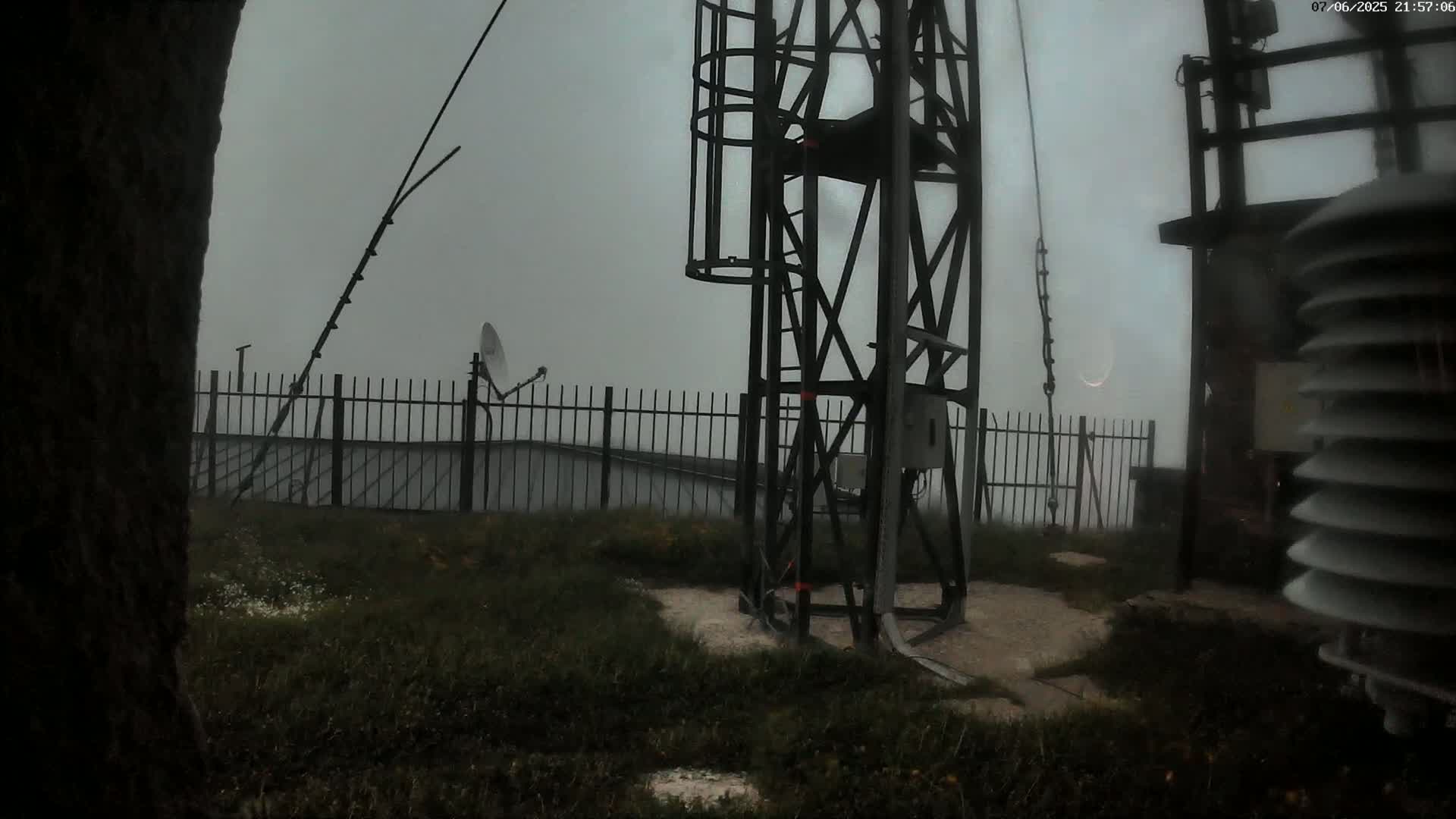 A metal tower with various equipment stands on a grassy area behind a metal fence, partially obscured by fog or mist.