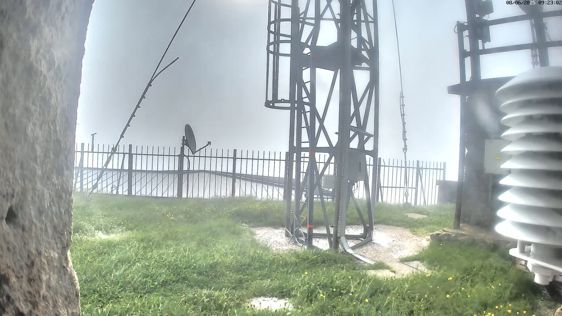 A metal tower with a satellite dish and weather monitoring equipment stands in foggy conditions, surrounded by short grass and a metal fence.