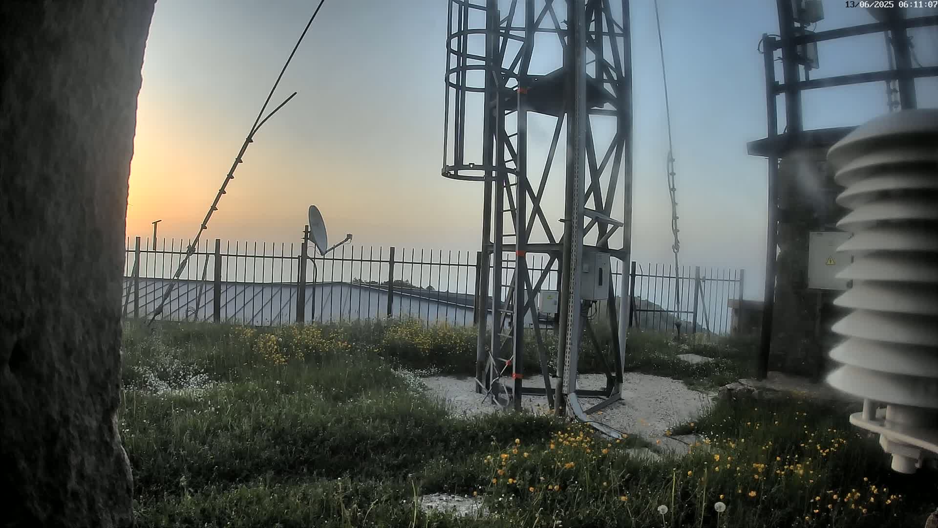 A metal tower with a satellite dish and weather station stands on a grassy hilltop at sunrise, behind a metal fence.