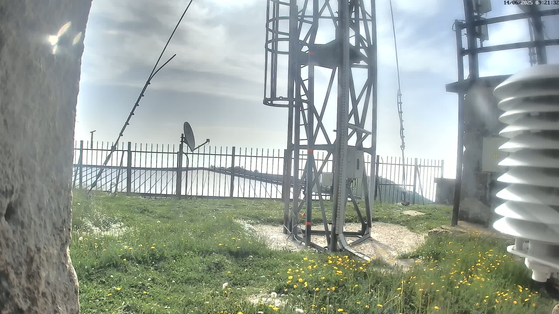 A metal tower with attached equipment stands on a grassy area behind a fence, under a cloudy sky.