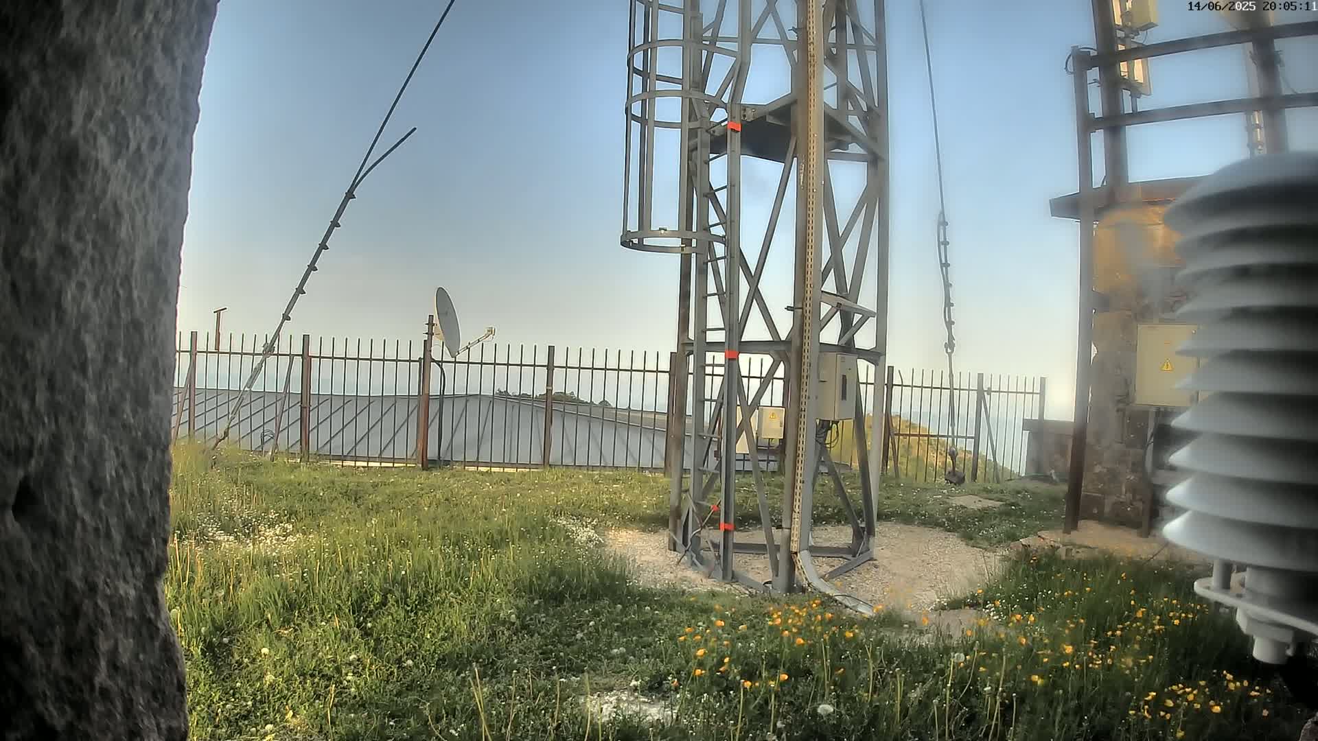 A metal communications tower stands in a grassy area with a fence, satellite dish, and weather station, under a clear, light-blue sky.