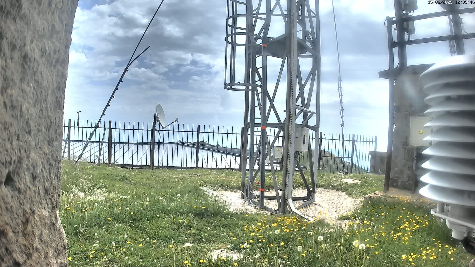 A metal tower with antennas and satellite dishes stands on a grassy hill under a partly cloudy sky.
