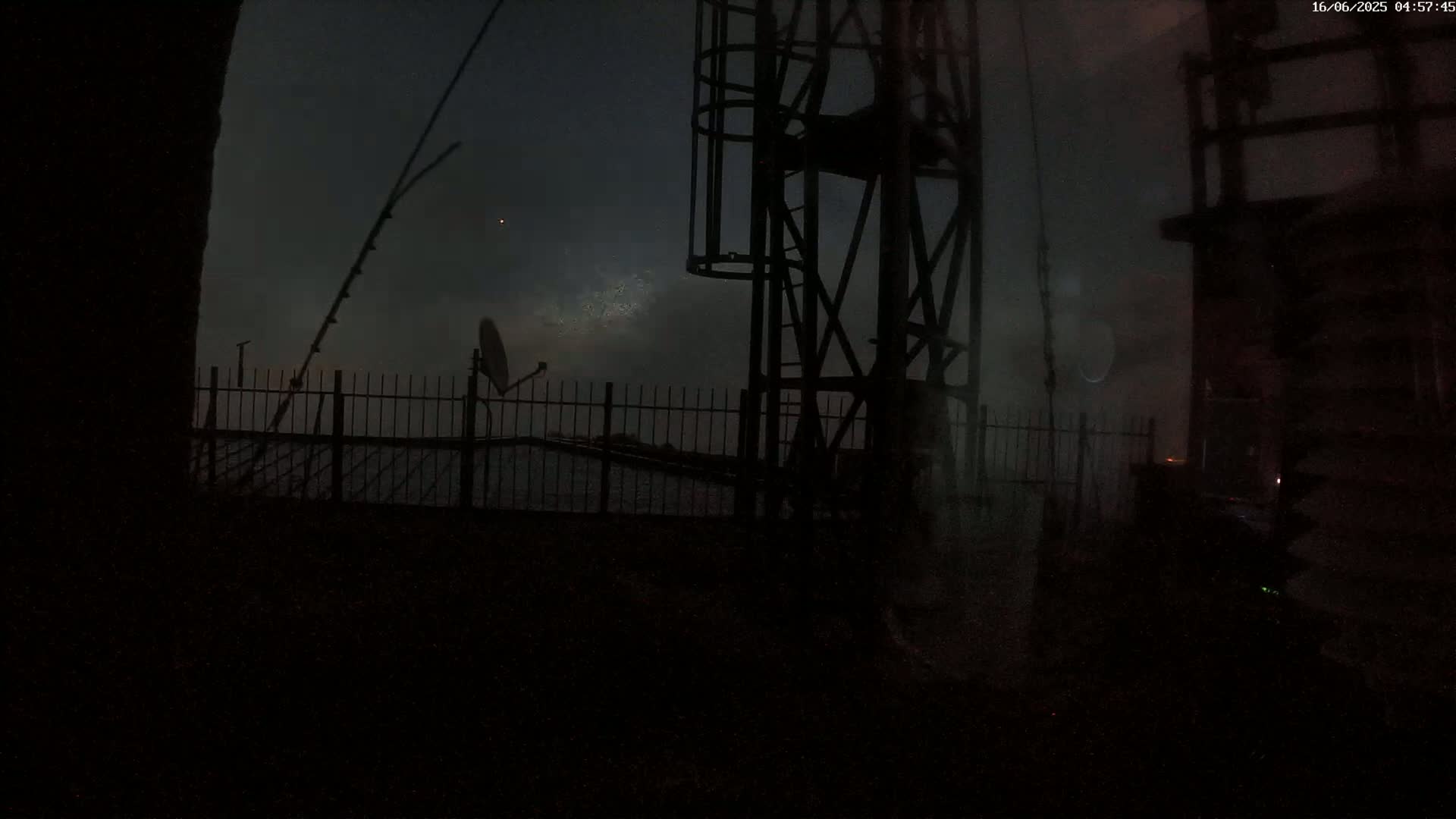 A dark, nighttime scene shows a metal fence, a satellite dish, and a tall metal structure under a misty, overcast sky.