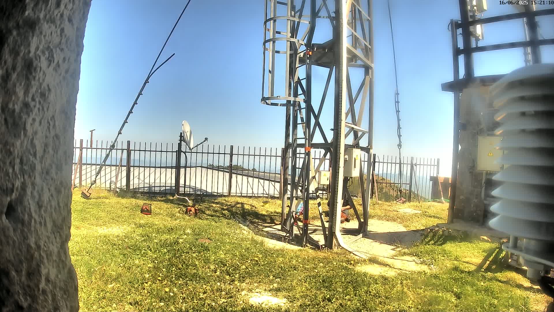 A metal tower with a ladder stands on a grassy area overlooking a calm ocean under a clear blue sky; nearby are some pieces of equipment and a satellite dish.