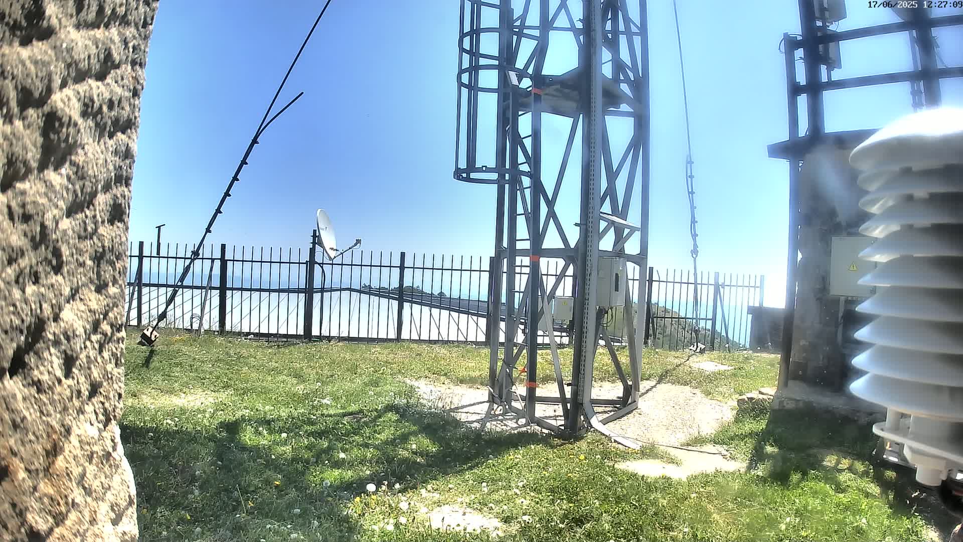 A tall metal tower with attached equipment stands on a grassy area with a fence behind it, overlooking a hazy ocean under a clear blue sky.
