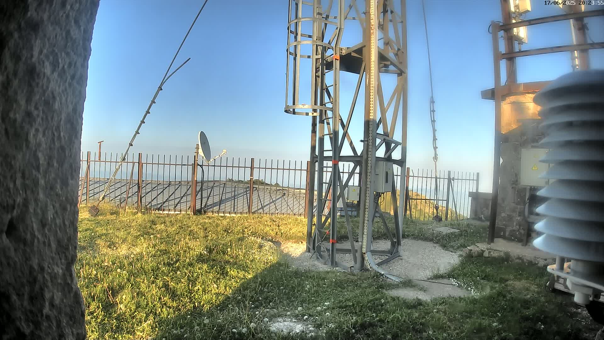 A tall metal tower with a ladder and attached equipment stands on a grassy hilltop under a clear blue sky, near a satellite dish and a weather station.