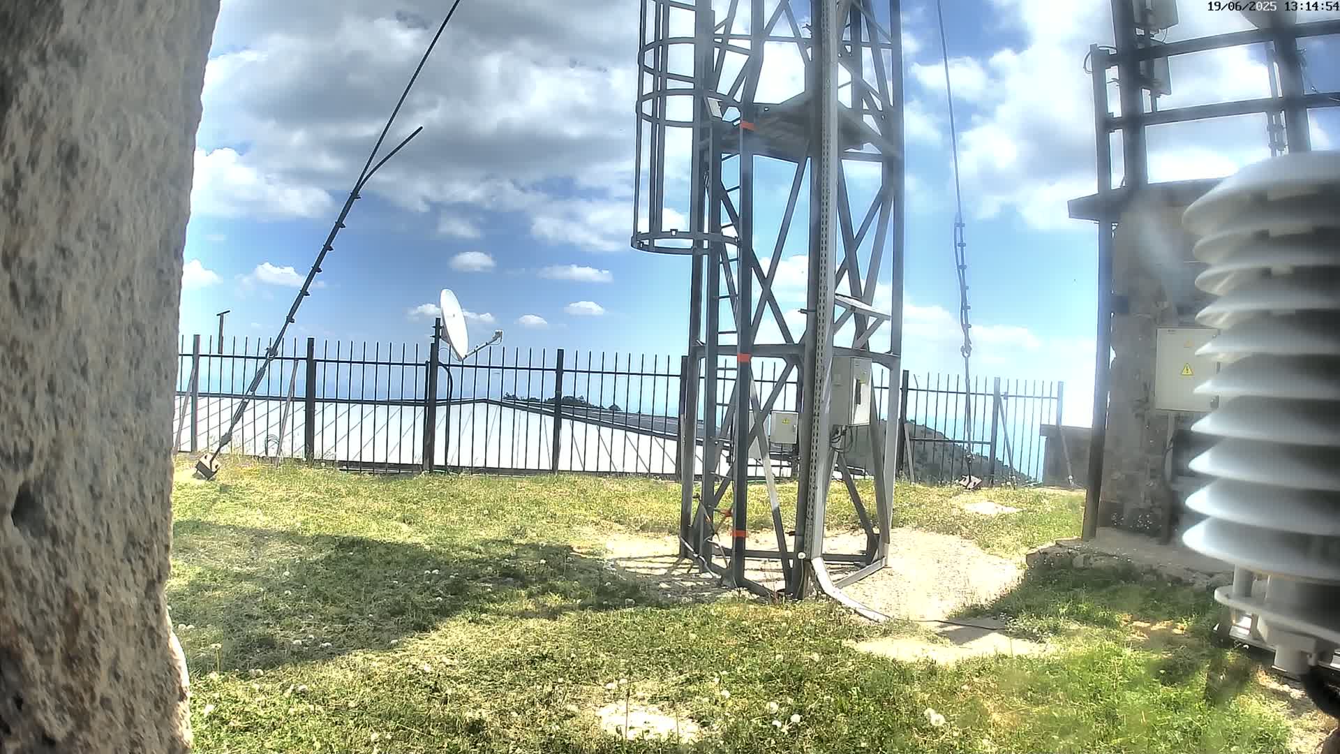 A metal tower with antennas and a satellite dish stands behind a black metal fence on a grassy area under a partly cloudy sky.