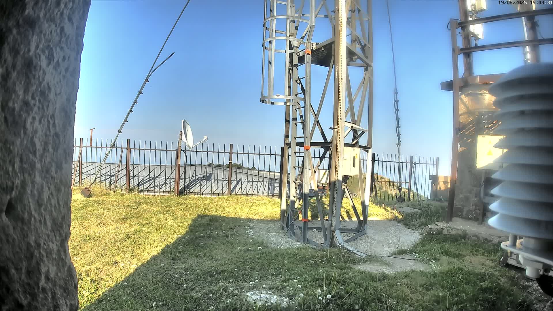 A metal tower with a ladder stands on grassy ground under a clear blue sky, near a fence, a satellite dish, and weather monitoring equipment.