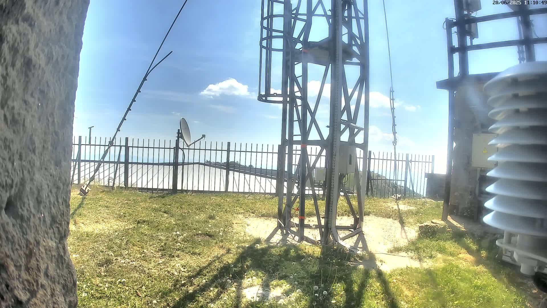 A metal tower with a satellite dish and other equipment stands on a grassy area behind a fence, under a partly cloudy sky.