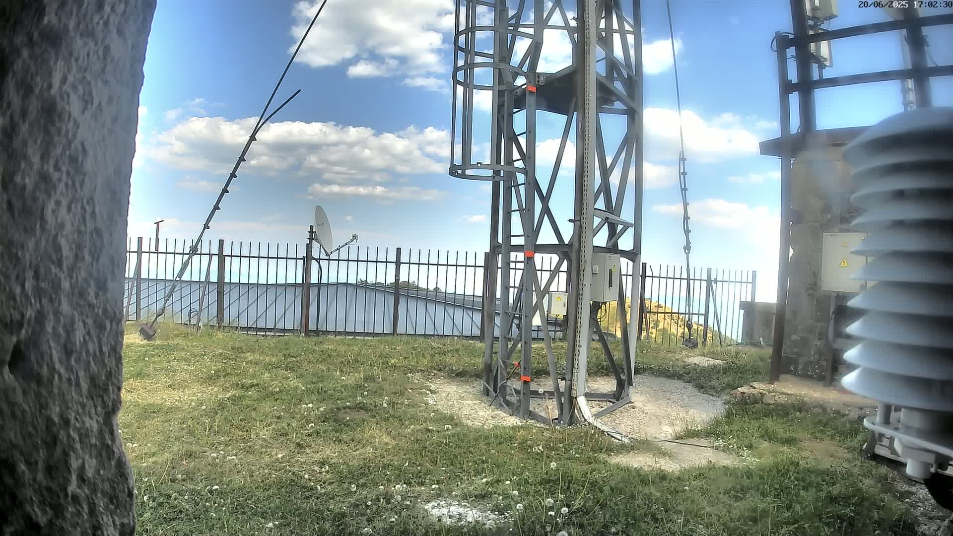 A tall metal tower with a satellite dish and weather equipment stands on a grassy area under a partly cloudy blue sky.