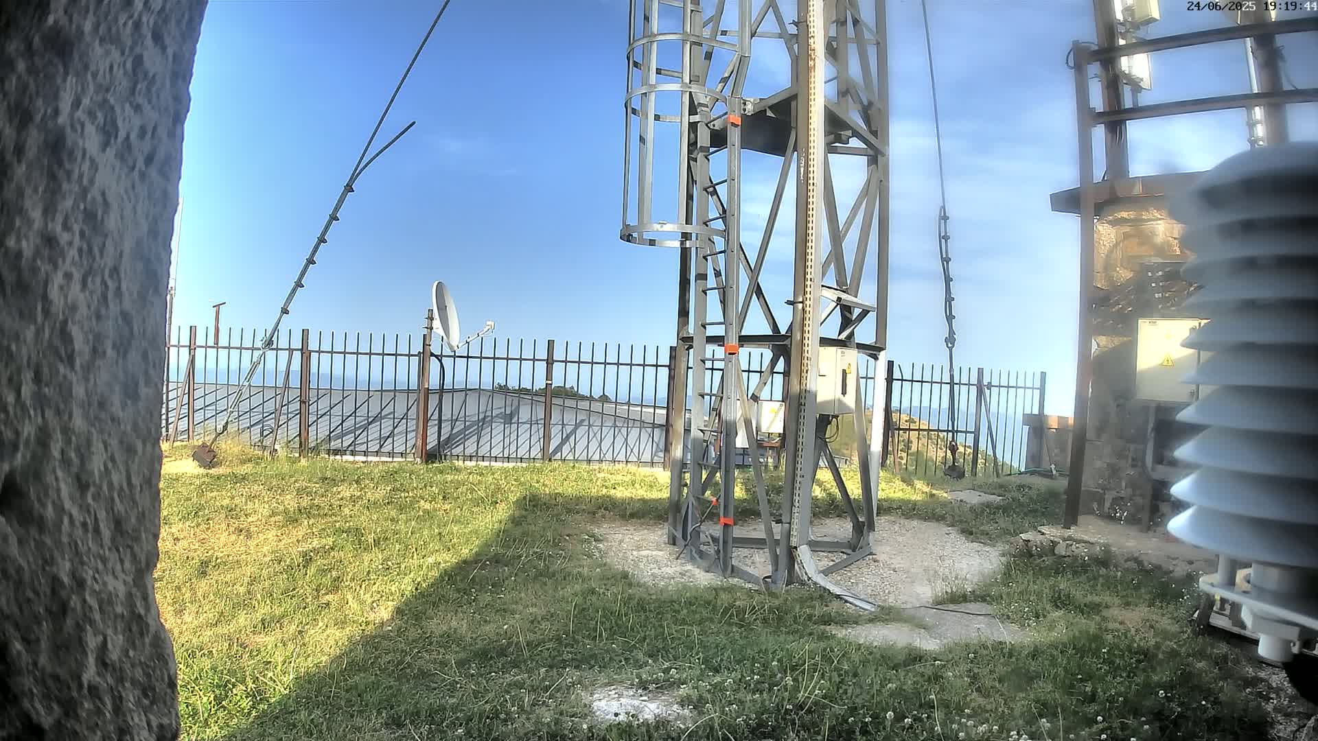 A metal tower with a ladder stands on grassy ground near a fence, a satellite dish, and other equipment, overlooking a body of water under a clear blue sky.