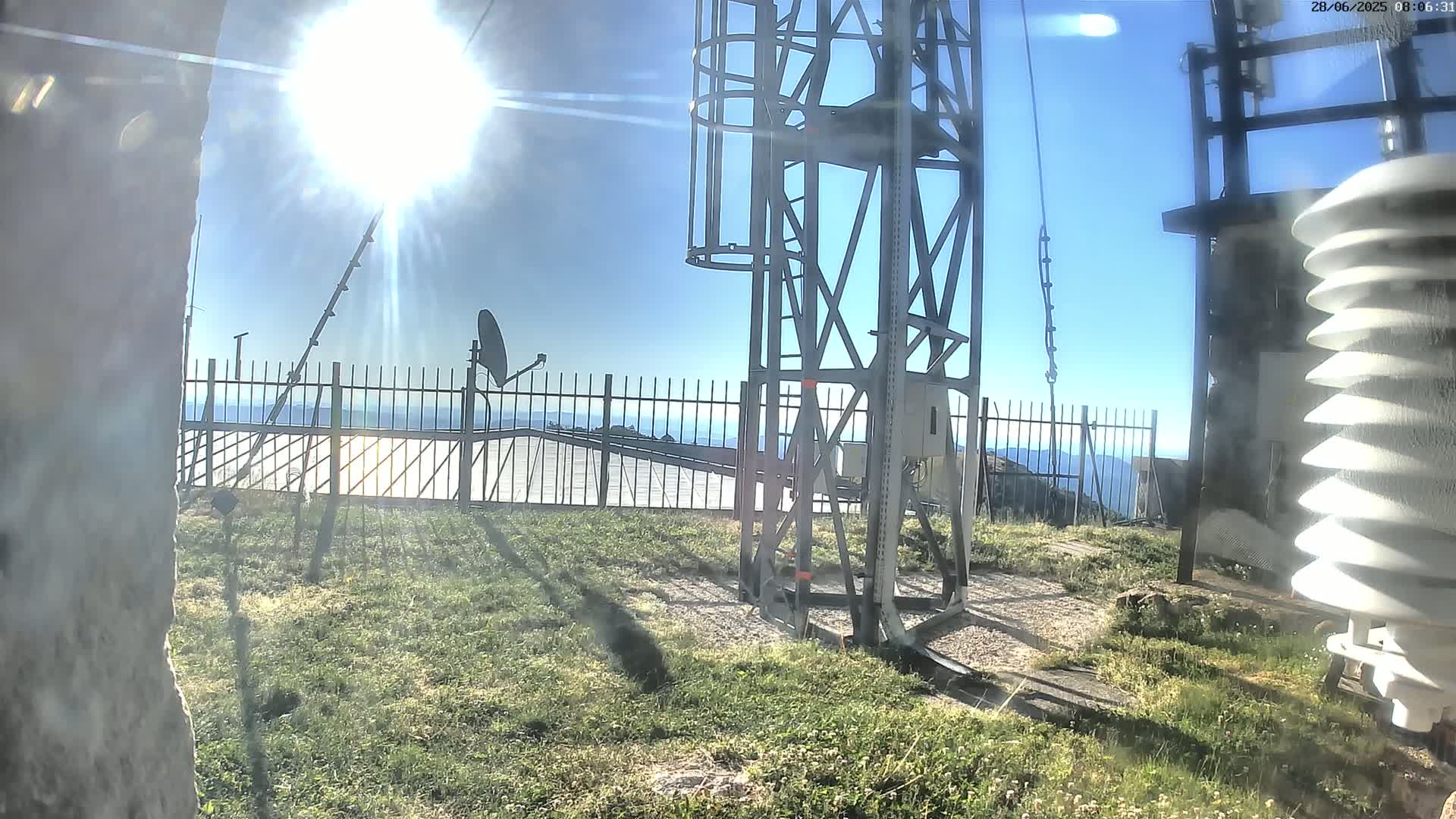 A sunny day reveals a metal tower and weather station on a grassy hilltop behind a metal fence, overlooking a distant valley.