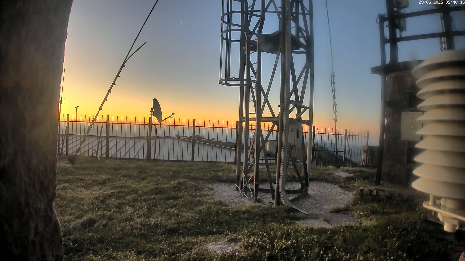 A metal communications tower stands on a grassy area overlooking a hazy sunrise over distant mountains.