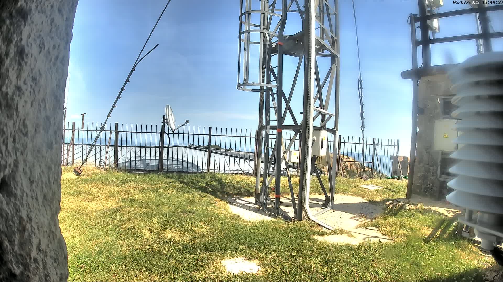 A metal tower with a ladder and various antennas stands on a grassy area near a fence overlooking a calm sea under a clear blue sky.