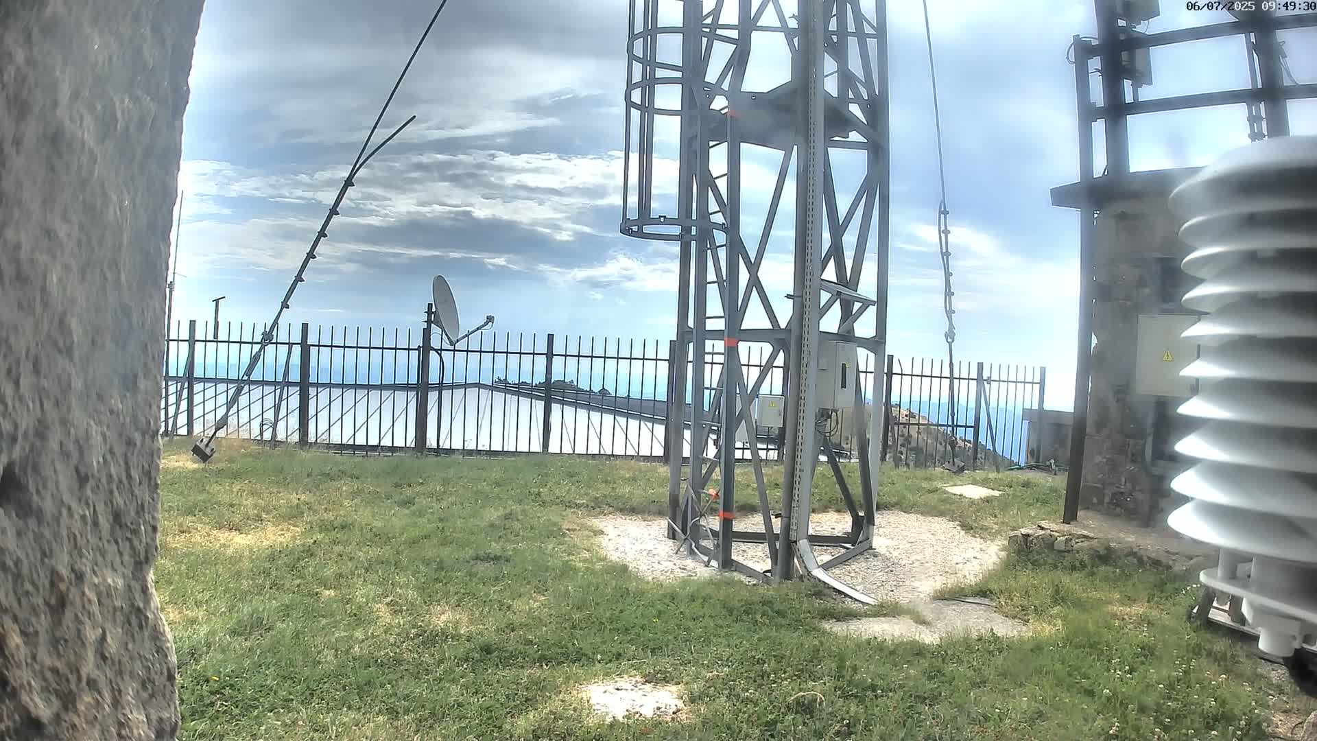 A metal tower with a ladder stands on a grassy area, with a satellite dish and a fence behind it, under a partly cloudy sky.