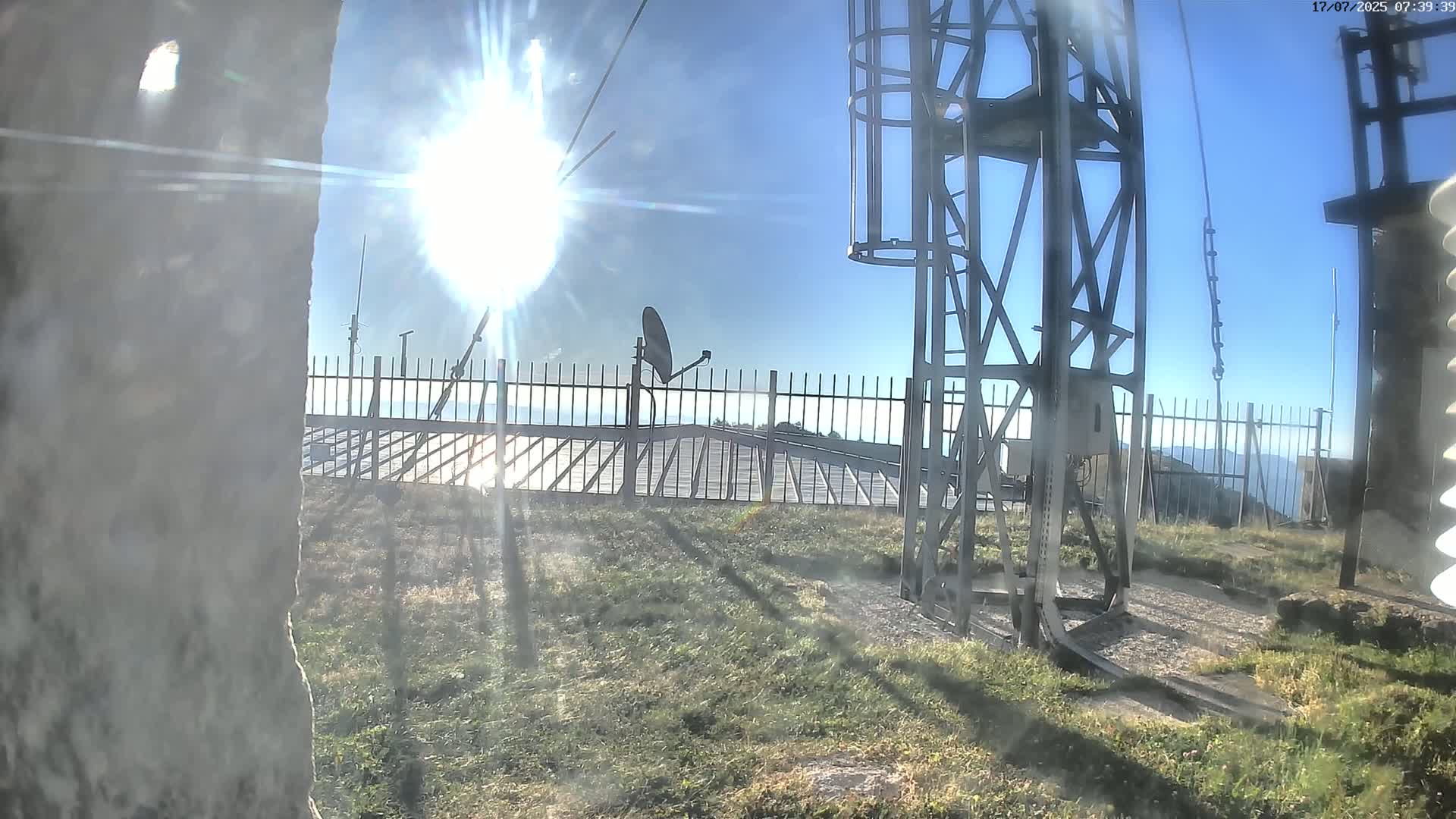A sunny day reveals a metal structure and satellite dish behind a fence on a grassy area overlooking a distant mountain range.
