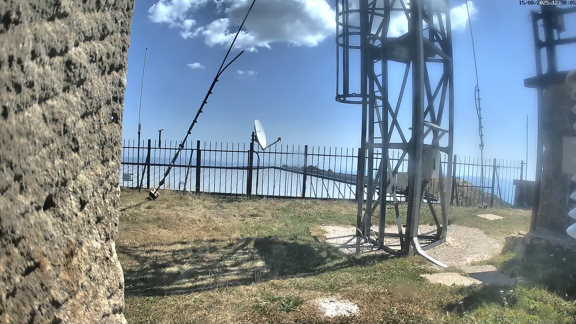 A sunny day reveals a metal structure and satellite dish behind a fence on a grassy area overlooking a distant mountain range.