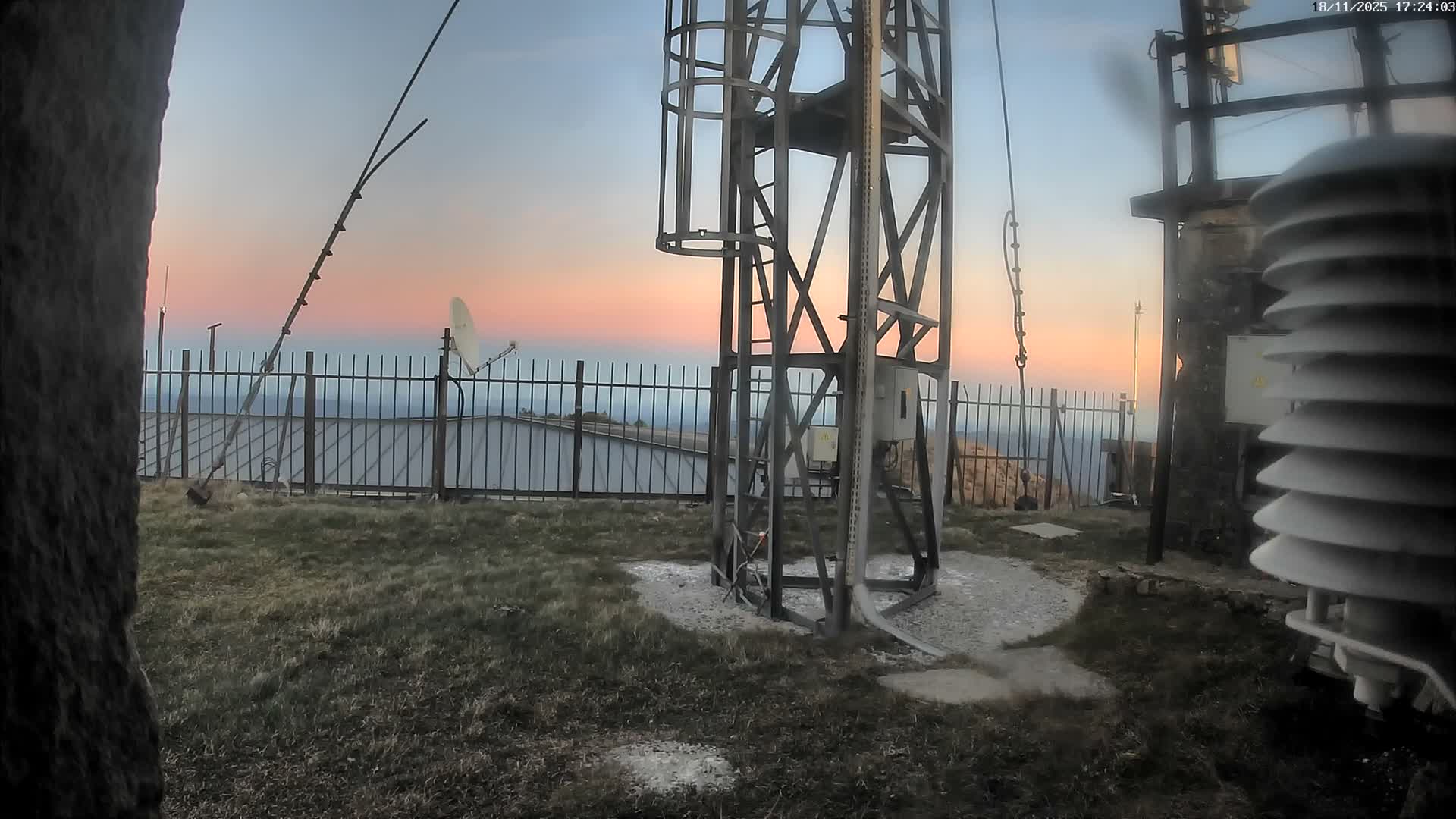 A sunny day reveals a metal structure and satellite dish behind a fence on a grassy area overlooking a distant mountain range.