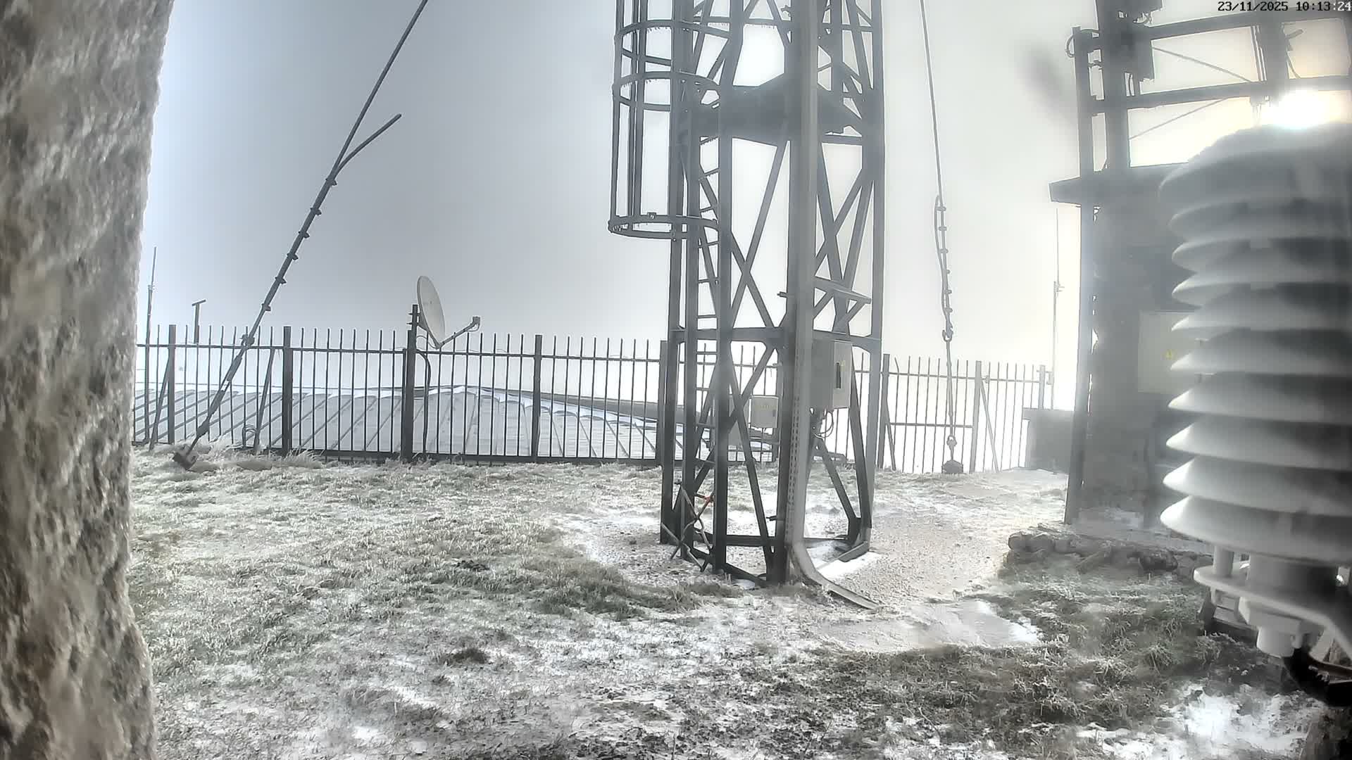 The image depicts a snowy and icy outdoor scene featuring a metal tower and various communication equipment, with patchy snow covering the ground under a foggy, overcast sky.