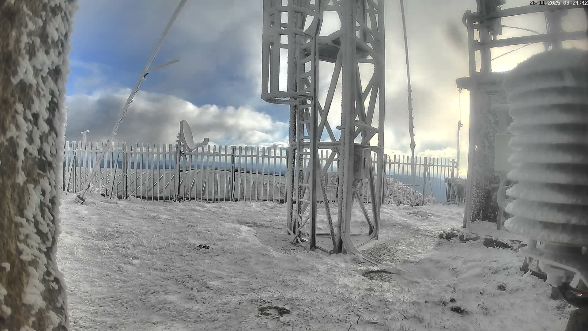An intensely cold, high-altitude scene reveals a metal communication tower, a fence, and ground entirely encased in rime ice and snow under a dramatic, partially cloudy winter sky.