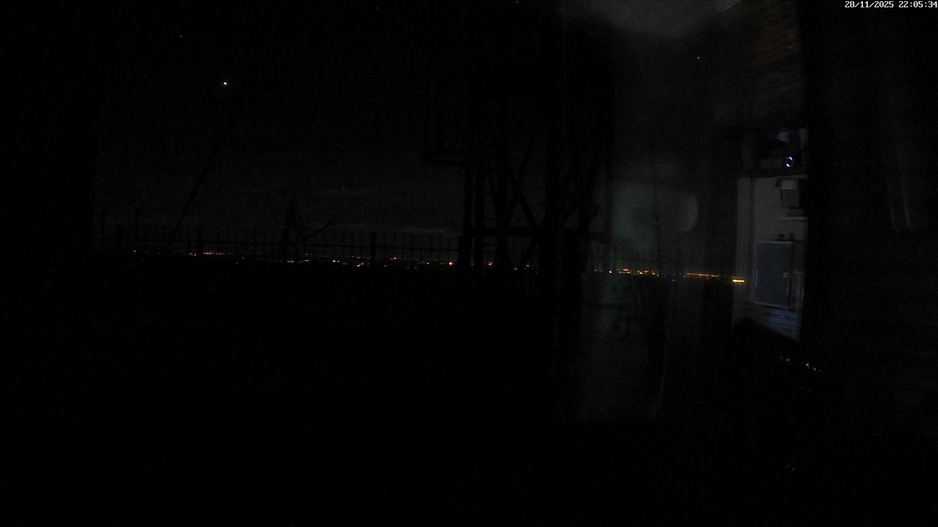 A dark, clear night view overlooks distant city lights from a high vantage point, with silhouetted structures and a fence in the foreground.
