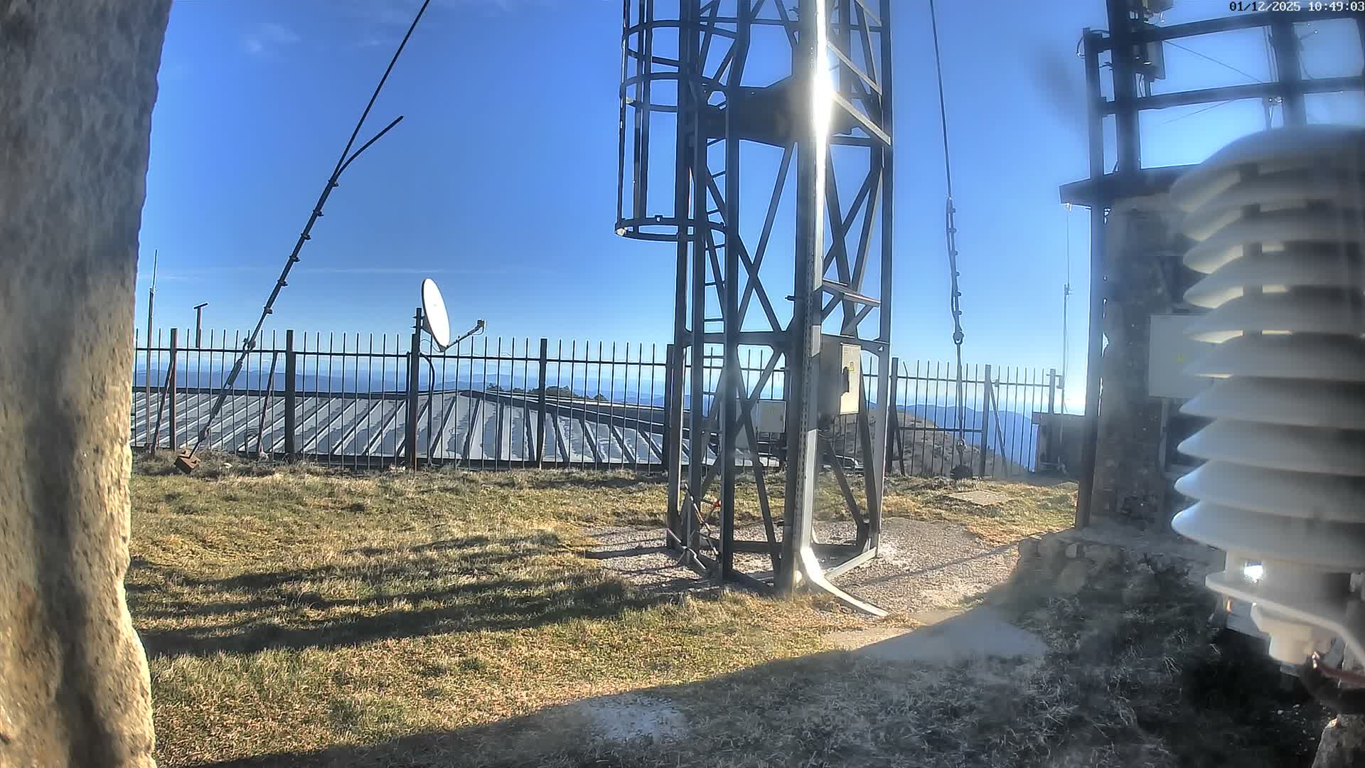 The image captures a sunny, clear day on a mountain peak, featuring a metallic communication tower and a white weather sensor in the foreground, with other antennas and a building visible behind a fence against a backdrop of distant hills and a bright blue sky.