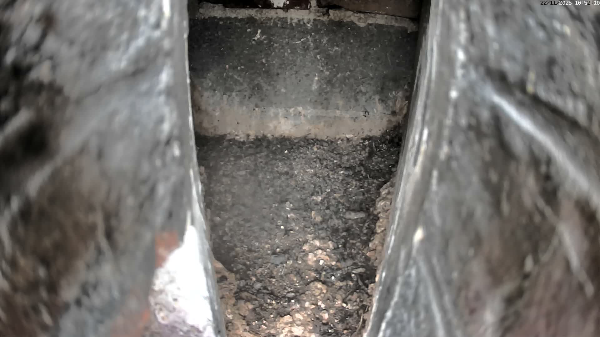 A dim, top-down view shows a narrow, enclosed channel between rough, dark walls, with its floor covered in dark granular debris and a horizontal lighter residue line visible on the back wall.