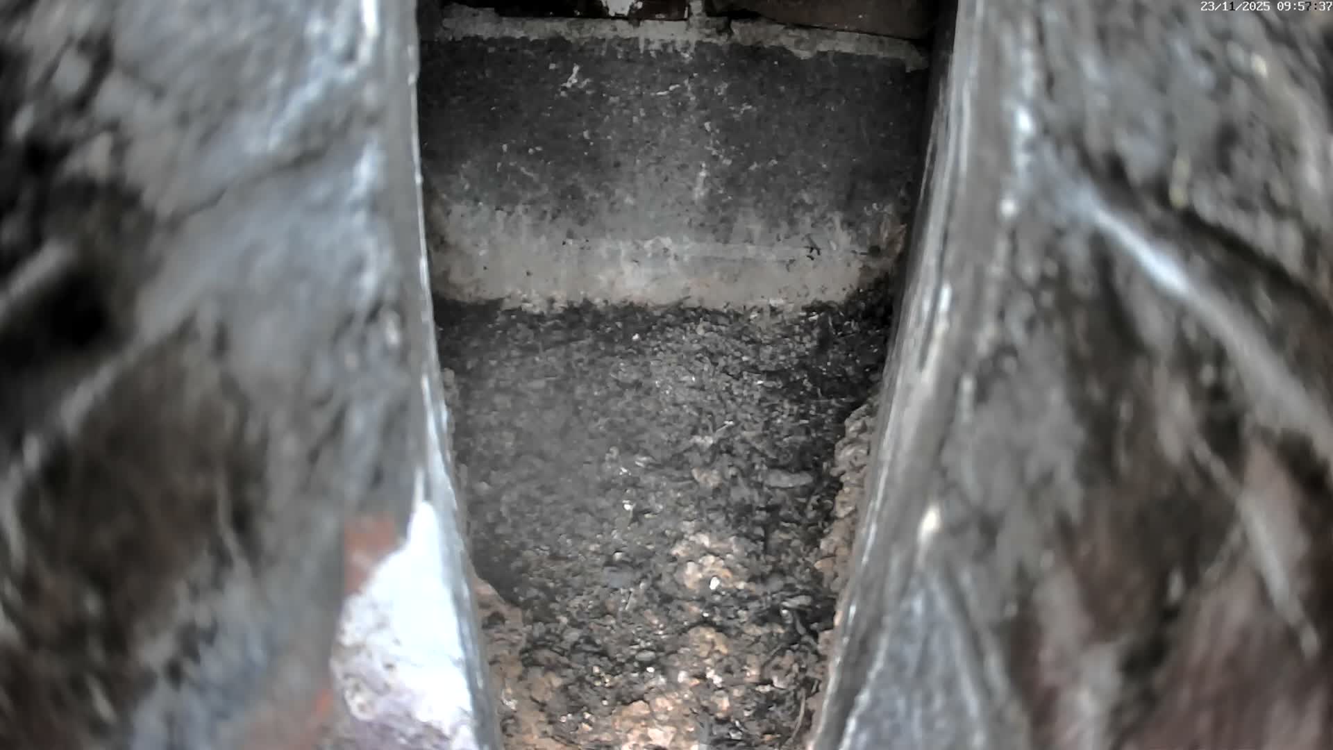 A dark, narrow cavity is seen from a downward perspective, revealing a base of granular debris beneath a lighter horizontal band, framed by blurry, dark textured surfaces.