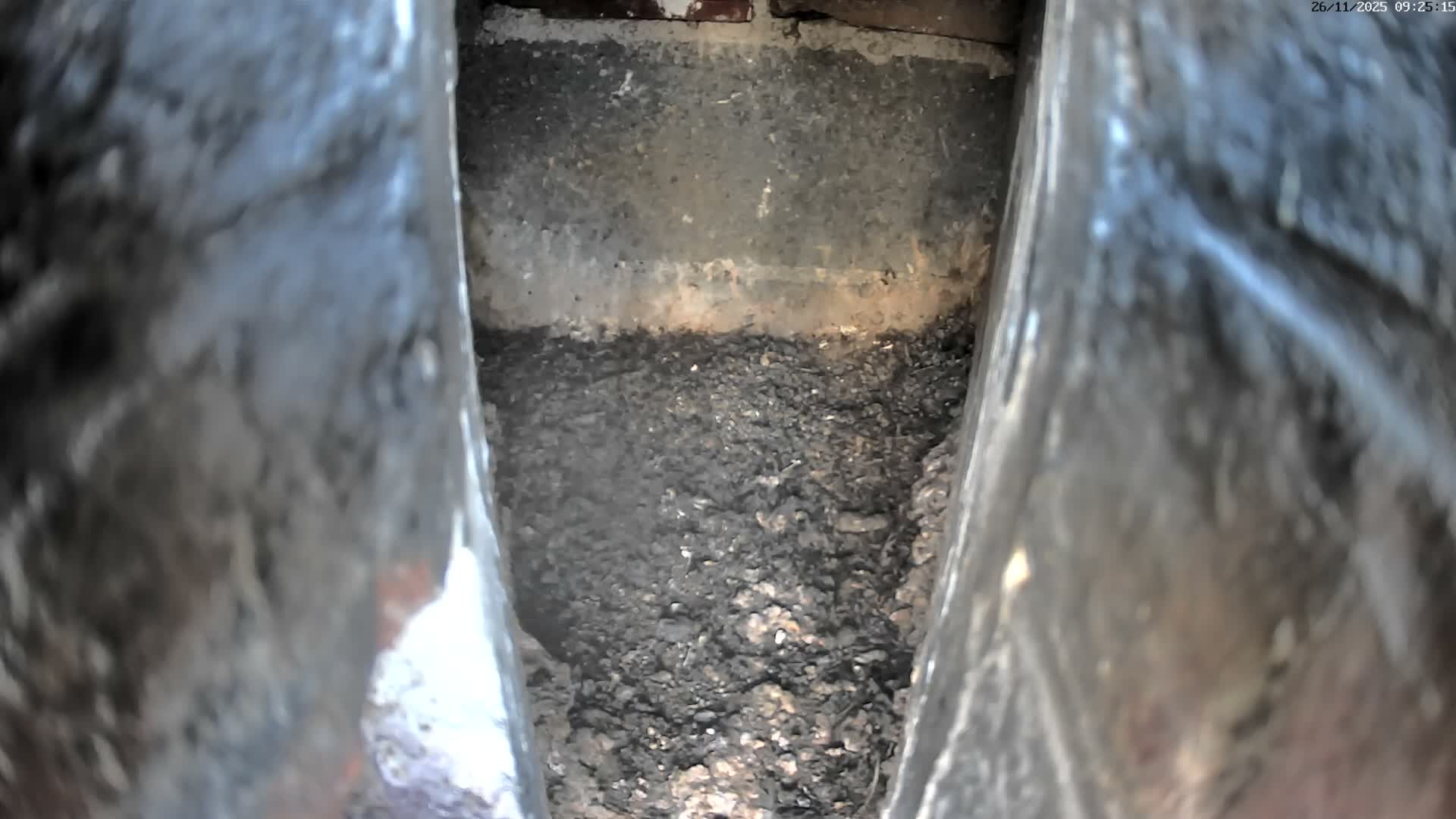 The image captures a dim, confined view between two blurry, dark vertical surfaces, revealing a rough back wall above a floor covered in dark, granular debris.