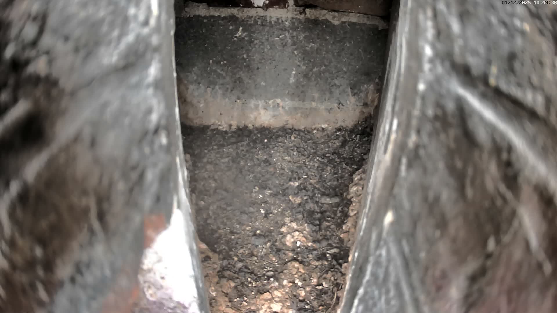 A downward view into a narrow, dimly lit interior shaft reveals a substantial accumulation of dark, granular debris at its bottom, framed by blurred, textured walls.