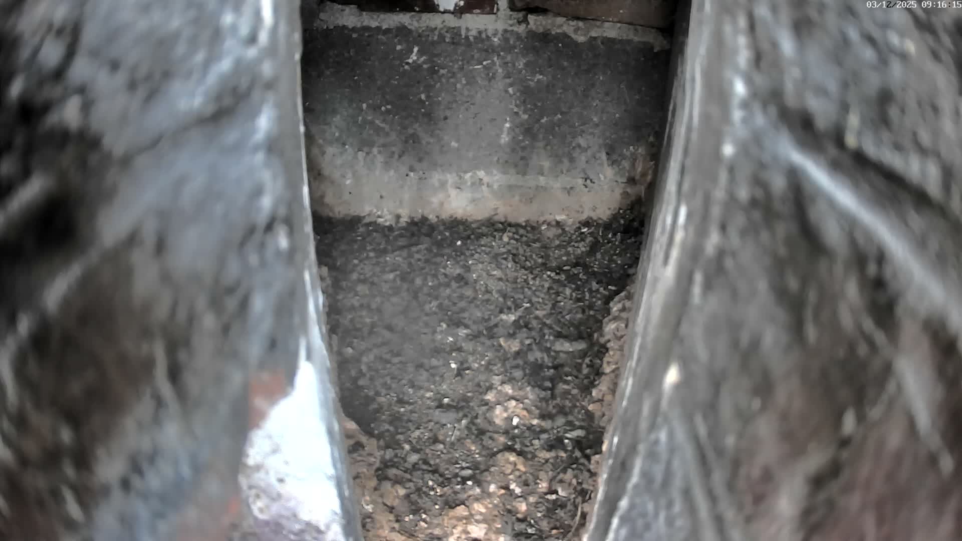 A narrow, dimly lit, downward view reveals a confined space with blurred, dark, textured walls on either side, a floor covered in dark granular debris, and a grimy back wall featuring a lighter horizontal band.