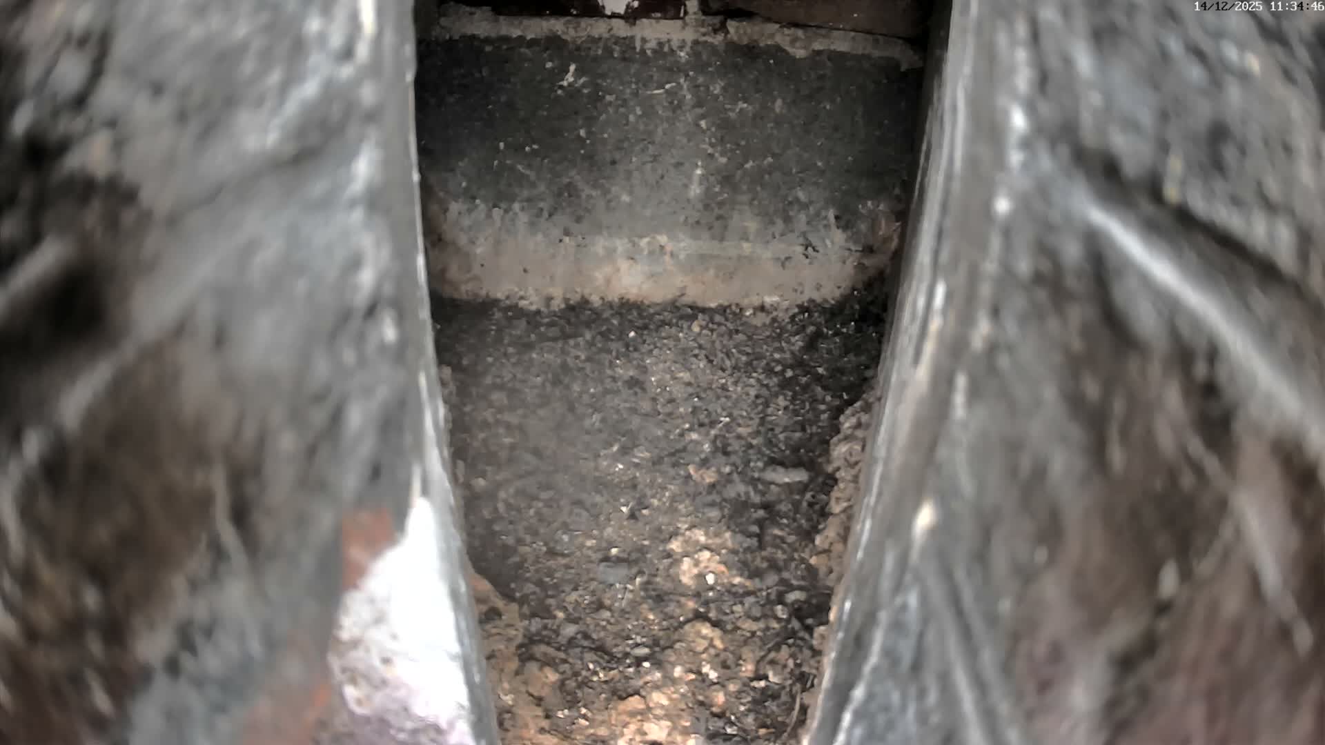 A narrow, dimly lit, downward view reveals a confined space with blurred, dark, textured walls on either side, a floor covered in dark granular debris, and a grimy back wall featuring a lighter horizontal band.