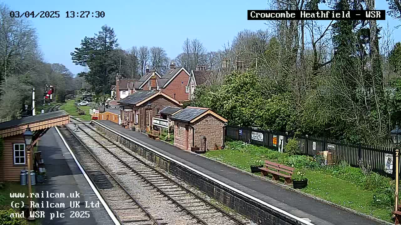 A sunny view of a train station with brick buildings, train tracks, and lush green vegetation surrounding it.