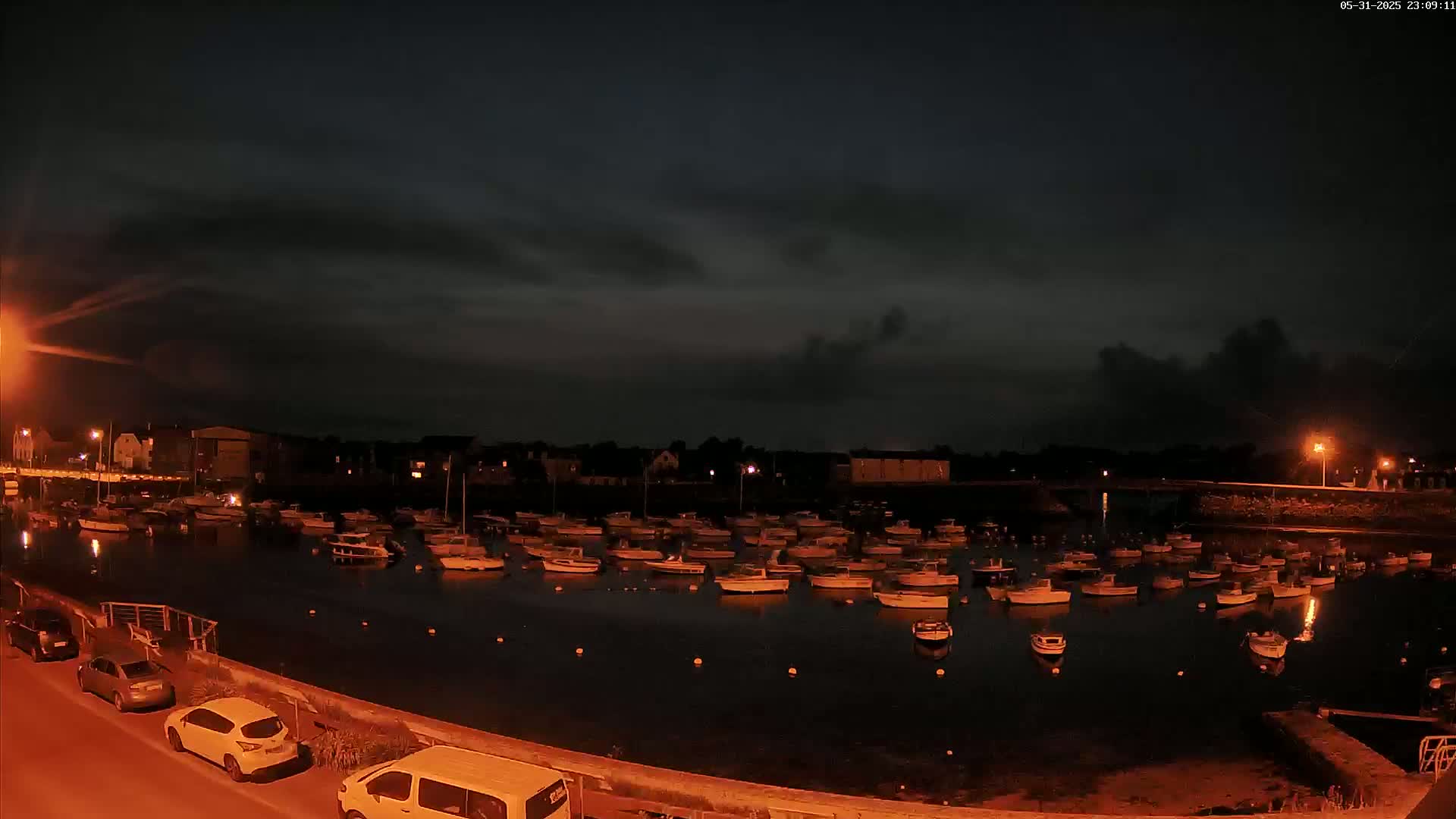 Under a dark, cloudy night sky, numerous small boats are moored in a calm harbor next to a waterfront lined with cars and buildings.