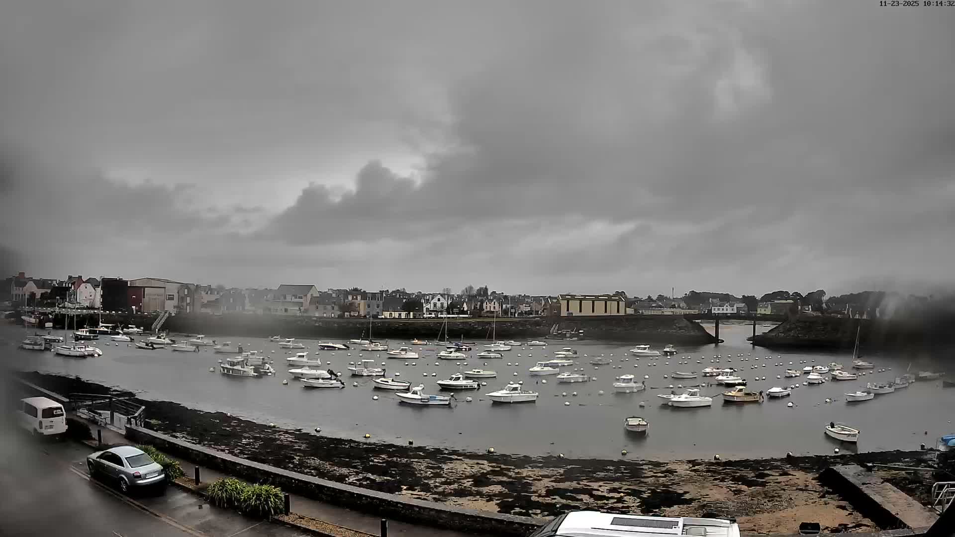 A broad bay densely populated with anchored boats is depicted on a heavily overcast and gloomy day, with a town lining the distant shore and a wet, dark foreground including a road and parked cars.