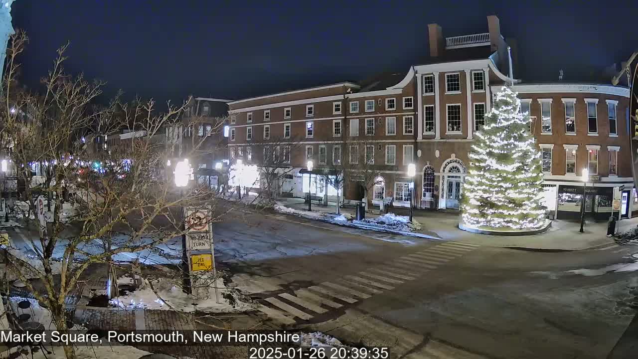 A snow-dusted town square at night features brick buildings, a large illuminated Christmas tree, and lightly trafficked streets.