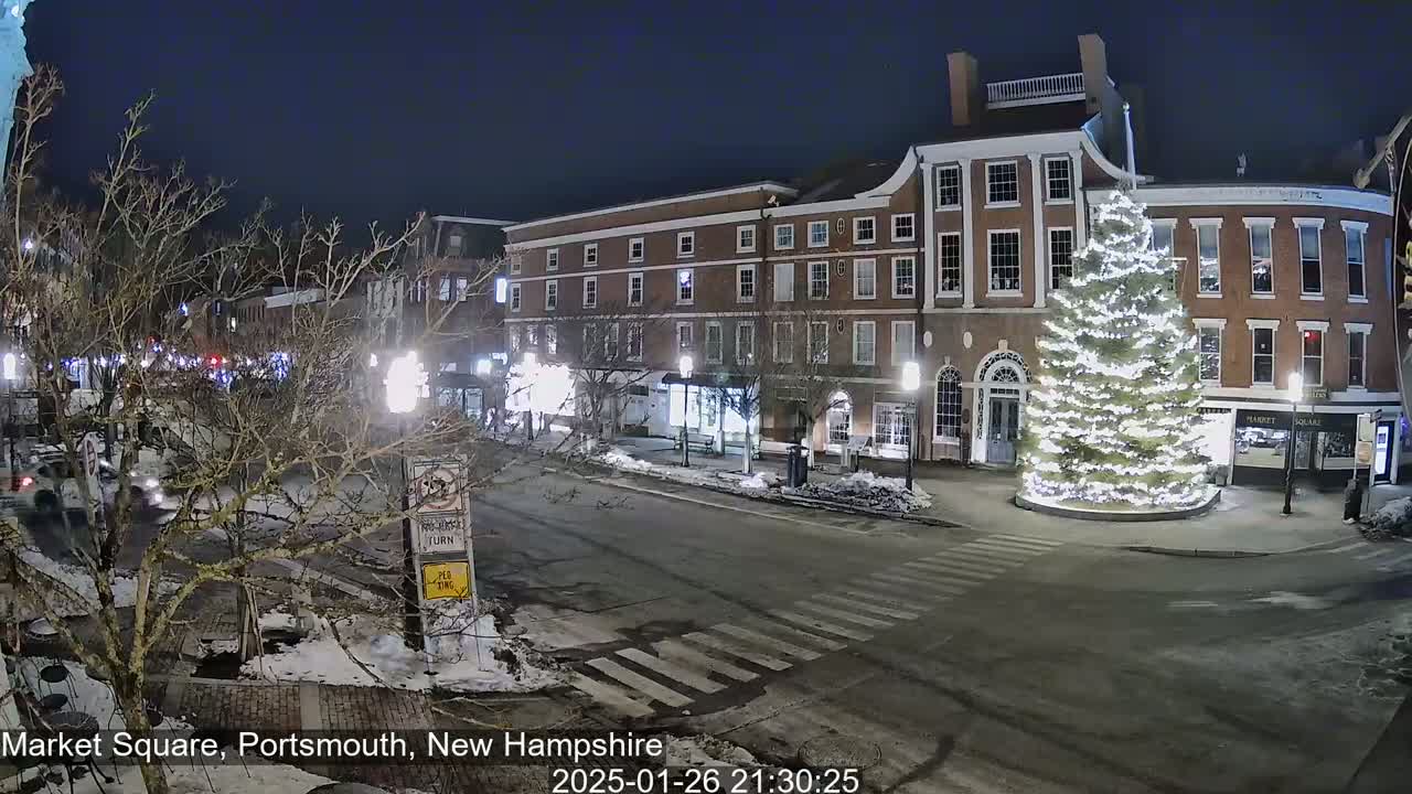 A nighttime view of a snow-covered Market Square in Portsmouth, New Hampshire, shows a large, illuminated Christmas tree in the center of the square, with brick buildings lining the street and a few cars visible.