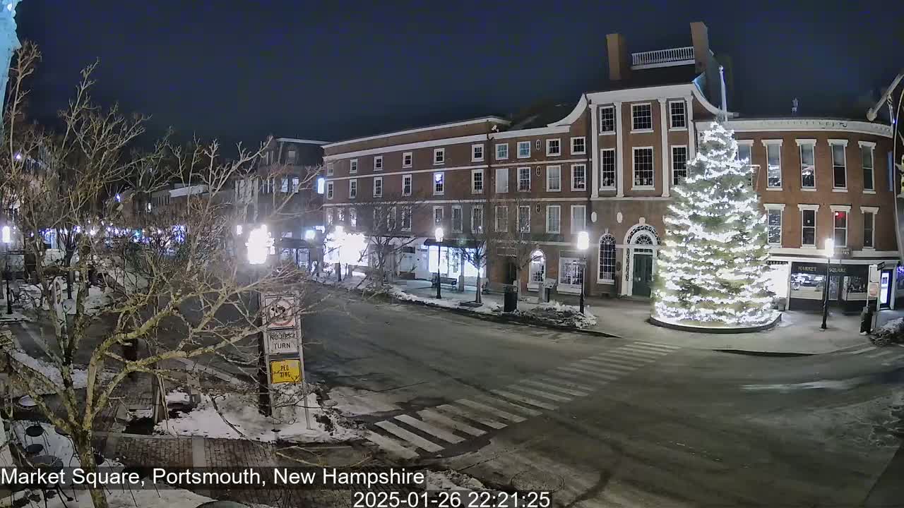 A snow-covered Market Square at night, illuminated by streetlights, features brick buildings and a large, brightly lit Christmas tree.