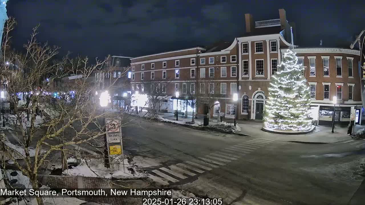 At night, a snow-dusted Market Square in Portsmouth, New Hampshire features a large, illuminated Christmas tree in the center of a street intersection, flanked by brick buildings and bare trees.