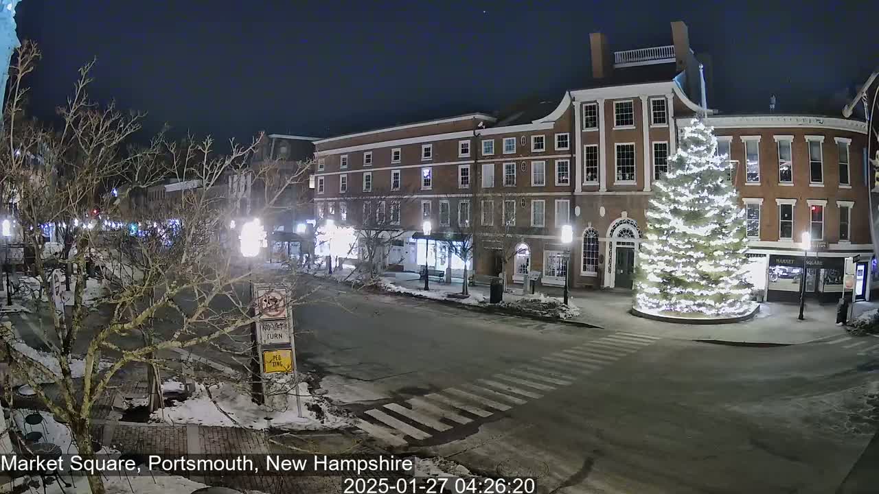 A snow-dusted, nighttime view of a town square with a large illuminated Christmas tree in the center, surrounded by brick buildings and bare trees.