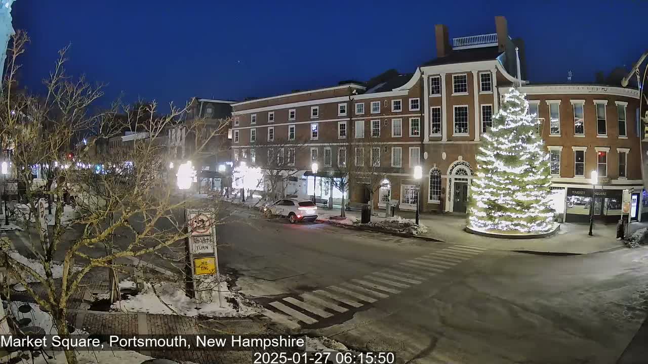 A snow-covered Market Square at night, illuminated by streetlights, features a large, brightly lit Christmas tree in the center, with brick buildings lining the square.