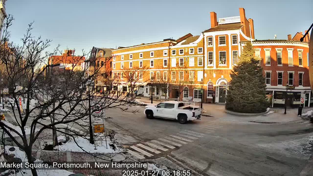 A white pickup truck drives through a snow-dusted town square, past brick buildings under a clear, sunny sky.