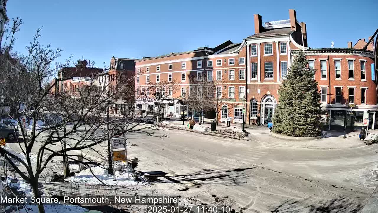A sunny day reveals a brick-lined town square with snow patches on the ground and bare trees, centered around a coniferous tree.